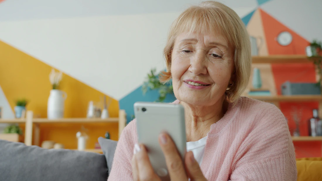 Uśmiechnięta starsza pani patrzy w telefon w kolorowym mieszkaniu